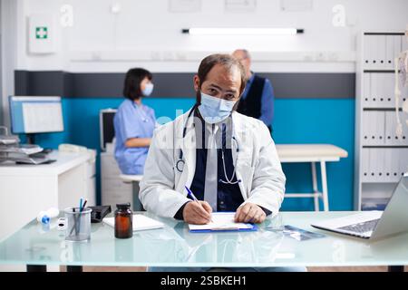 Der junge Arzt schreibt auf die Zwischenablage, während die Krankenschwester dem Rentner hilft, der im Hintergrund des klinischen Büros auf dem Krankenhausbett sitzt. Männlicher Arzt, der in die Kamera schaut und medizinische Aufzeichnungen am Schreibtisch überprüft. Stockfoto