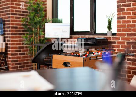 Selektiver Fokus auf Tischkicker, der in modernen gemauerten Arbeitsbereichen platziert wird und für Mitarbeiter bereit ist, sich zu entspannen. Leeres Startup-Büro mit Computer, Drucker, Pflanzen und Pausenbereich zur Entspannung. Stockfoto