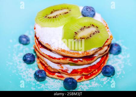Stapel Pfannkuchen mit Puderzucker und umgeben von Heidelbeeren auf einer hellblauen Oberfläche. Pfannkuchen mit Sahne, Kiwi-Scheiben Stockfoto