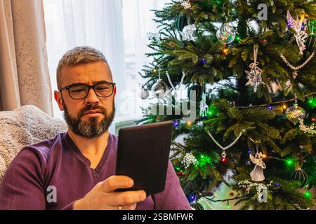 Ein junger Mann entspannt sich in einem bequemen Stuhl, vertieft in das Lesen eines E-Buches, während festliche Lichter auf dem Weihnachtsbaum in der Nähe funkeln und eine warme Atmosphäre schaffen Stockfoto