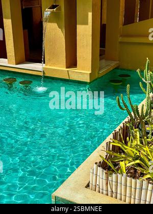 Tropischer Pool mit Pflanzen und Sunny Modern Architecture Ein sonniger türkisfarbener Pool umgeben von moderner gelber Architektur und tropischen Pflanzen Stockfoto