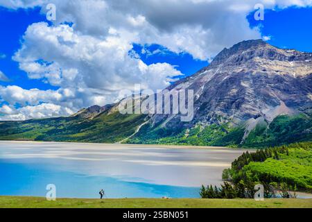 Der Mann steht auf einem grasbewachsenen Hügel mit Blick auf einen See. Der Himmel ist bewölkt und die Berge im Hintergrund sind mit Schnee bedeckt Stockfoto