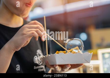 Zugeschnittene Aufnahme einer glücklichen jungen, erwachsenen asiatischen Foodie-Frau, die auf dem Markt gebratene Schnittlauch hält und isst. Street-Food-Konzept Stockfoto