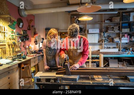 Anfängerin Frau Uhr Meister Zimmermann bedienen Säge Maschine in Werkstatt. Ehemann und Ehefrau arbeiten zusammen Stockfoto