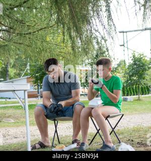 Ein Mann und ein Junge sitzen an einem kleinen Klapptisch in einem Park, tragen schwarze Handschuhe, während sie aus Einwegbehältern essen. Sie sind entspannt und genießen ihre Stockfoto