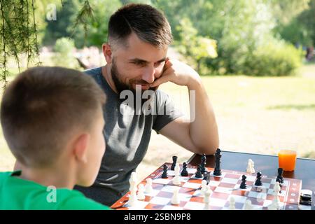 Ein Mann und ein Junge sind vertieft in ein Schachspiel an einem Tisch im Freien. Die Szene ist ruhig und idyllisch, in Sonnenlicht getaucht. Stockfoto