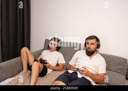 Ein Mann und ein Junge, die Kopfhörer tragen, sitzen bequem auf einer Couch und konzentrieren sich auf ihre eigenen tragbaren Videospielkonsolen. Eine gemütliche, entspannte Szene. Stockfoto