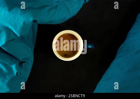 Kaffeetasse im Bett auf dunkelschwarzem und blaublauem Laken mit kleinem Herz im Schaum, Blick von oben Stockfoto