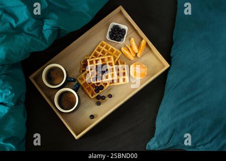 Waffeln, Kaffee, Blaubeeren und Orangenfrühstück auf einem Holztablett im Bett auf schwarzen und blaublauen Laken, Blick von oben Stockfoto