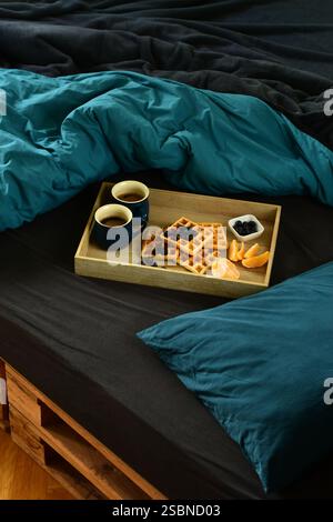 Waffeln, Kaffee, Blaubeeren und Orangenfrühstück auf einem Tablett im Holzpalettenbett auf schwarzen und dunkelblauem Laken Stockfoto