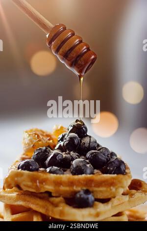 Nahaufnahme von Honig, der aus einem hölzernen Honiglöffel über einem Stapel belgischer Waffeln mit Heidelbeeren drauf tropft, mit Bokeh-Lichtern im Hintergrund Stockfoto