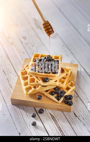 Honig tropft aus einem Honiglöffel auf einem Stapel belgischer Waffeln mit Heidelbeeren in der Morgensonne auf einem Holztisch Stockfoto