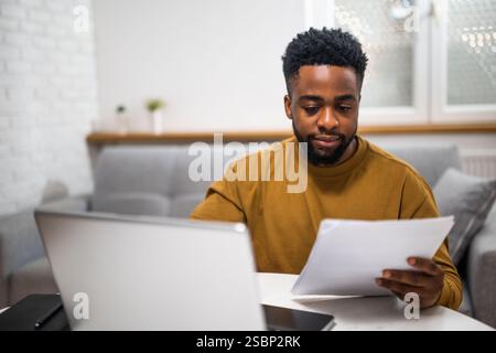 Erfolgreicher afroamerikanischer Geschäftsmann, der bei der Arbeit von zu Hause aus ein Notebook verwendet und Dokumente analysiert. Stockfoto