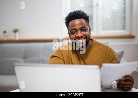 Porträt eines lächelnden jungen schwarzen Geschäftsmannes, der sein Notebook benutzt und Dokumente untersucht, während er in seinem gemütlichen Zuhause arbeitet. Stockfoto