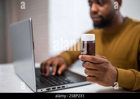 Nahaufnahme eines schwarzen Mannes, der die Pille in der Hand hält und einen Laptop benutzt, um mehr Informationen über sein verschreibungspflichtiges Medikament zu suchen. Stockfoto
