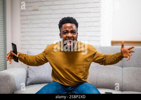 Enttäuschter junger afroamerikanischer Mann, der schreiend und wütende Gesten macht, während er Fußballspiele im fernsehen bei sich zu Hause ansieht. Stockfoto
