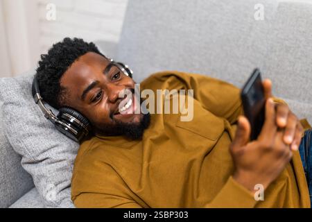 Fröhlicher junger schwarzer Mann genießt die Verwendung von schnurlosen Kopfhörern und Mobiltelefonen, während er in seinem gemütlichen Zuhause auf dem Sofa liegt. Stockfoto