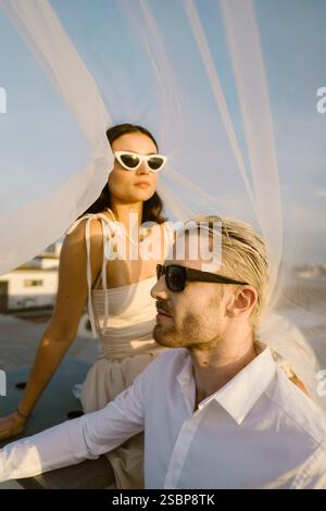 Ein junges verheiratetes Paar trägt eine Sonnenbrille, während es unter dem Schleier auf dem Dach sitzt Stockfoto