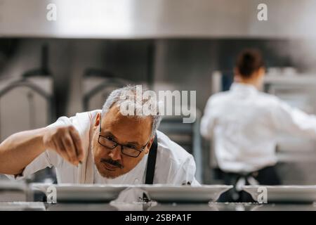 Reifer männlicher Koch in Brillen garniert Essen während der Arbeit in der kommerziellen Küche Stockfoto