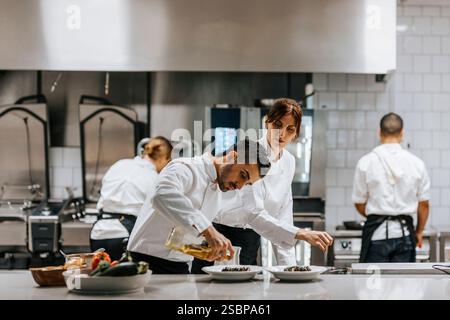 Männlicher Koch gießt Öl auf das Gericht, während weibliche Mitarbeiterin in der Nähe der Küchenzeile im Restaurant steht Stockfoto