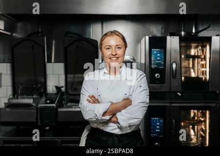 Porträt einer lächelnden Küchenchefin in weißer Uniform und stehend mit überkreuzten Armen in der Küche im Restaurant Stockfoto