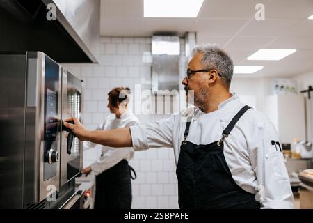 Reifer männlicher Koch, der Schürze mit Backofen in der kommerziellen Küche trägt Stockfoto