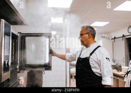 Reifer männlicher Koch, der den Ofen öffnet, während er in der kommerziellen Küche steht Stockfoto