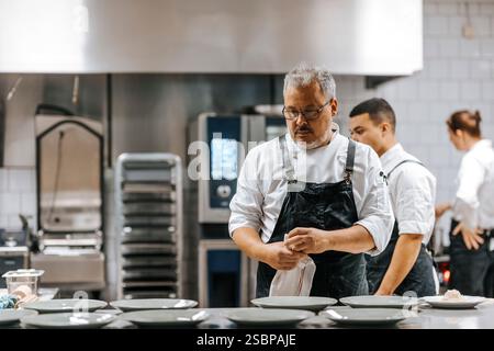 Reifer männlicher Koch mit schwarzer Schürze und Blick auf leere Teller auf der Küchentheke im Restaurant Stockfoto
