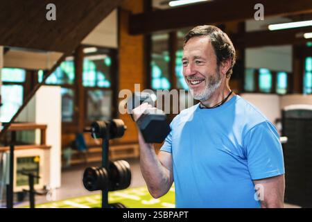 Ein Mann lächelt, während er eine Kurzhantel hebt. Er ist in einem Fitnessstudio und genießt sein Training Stockfoto