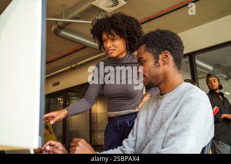 Niedriger Blickwinkel eines jungen Programmierers, der mit einem männlichen Kollegen spricht, der im Büro am Desktop-PC arbeitet Stockfoto