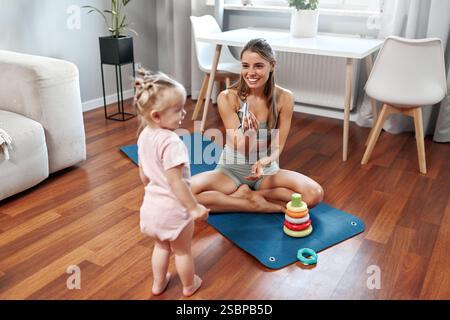 Eine fröhliche und fröhliche Mutter übt ihre Yoga-Routine zu Hause, während ihr liebenswertes Baby in der Nähe spielt und eine wunderbare Mischung aus Passform bietet Stockfoto