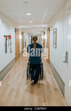 Rückansicht einer Pflegeperson, die älteren Patienten im Rollstuhl hilft, während sie in einem Pflegeheim im Korridor laufen Stockfoto