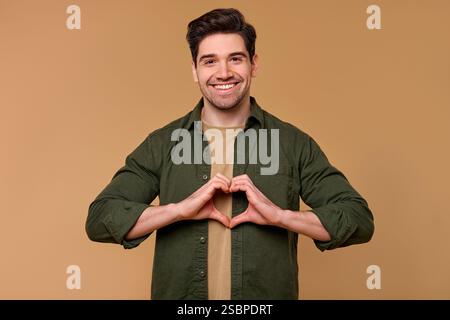 Ein fröhlicher junger Mann, der Glück und Liebe ausstrahlt, lächelt hell und kreiert mit seinen Händen eine Herzform. Sein fröhlicher Ausdruck verkörpert po Stockfoto