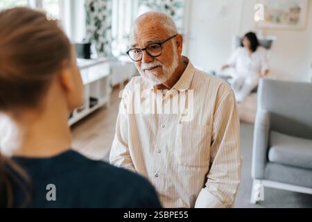 Pensionierter Seniorenmann, der mit einer Betreuerin spricht, während er im Pflegeheim sitzt Stockfoto