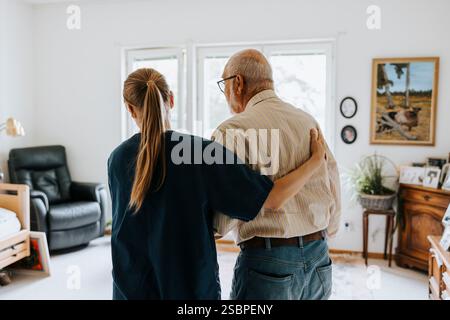 Rückansicht einer Krankenschwesterin, die einen älteren Mann beim Gehen im Altenpflegeheim unterstützt Stockfoto