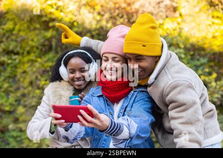 Drei glückliche Freunde machen im Herbst ein Selfie in einem Park, einer zeigt auf etwas in der Ferne Stockfoto