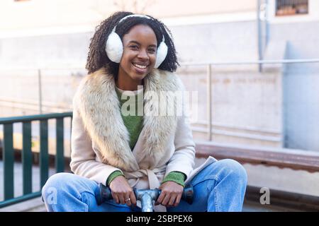 Porträt einer jungen schwarzen Frau, die Ohrenschützer und Winterkleidung trägt, lächelt und in der Stadt auf einer Schaukel sitzt Stockfoto