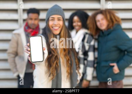 Lächelnde junge Frau, die ihr Smartphone mit weißem, leerem Bildschirm zeigt und Winterkleidung trägt, mit multiethnischen Freunden im Hintergrund außer Fokus steht Stockfoto