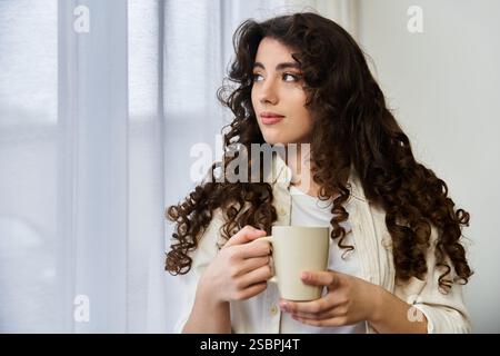 Eine junge Frau sitzt allein zu Hause, schlürft an ihrem warmen Getränk und blickt nach draußen. Stockfoto