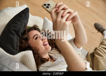 Die schöne junge Frau liegt auf einer gemütlichen Couch und schaut zuhause glücklich auf ihr Smartphone. Stockfoto