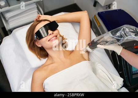 Eine schöne junge Frau lächelt, während sie in einer schlanken Klinik eine Laser-Haarentfernungsbehandlung erhält. Stockfoto