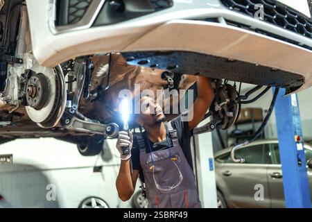 Professioneller Mechaniker mit Taschenlampe, der die Unterseite des angehobenen Fahrzeugs in einer gut ausgestatteten Autowerkstatt untersucht Stockfoto