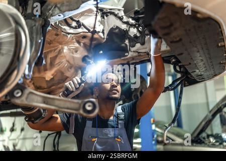 Junger Mechaniker, der die Taschenlampe hält, um das Fahrgestell während der Wartung in einer professionellen Werkstatt zu überprüfen Stockfoto