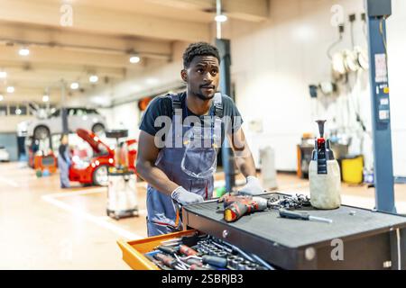 Ein junger afrikanischer Automechaniker schiebt einen Werkzeugwagen in einer geschäftigen Werkstatt, um ein Auto zu reparieren Stockfoto