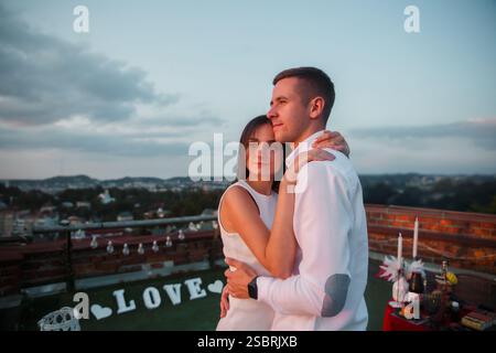Ein Paar steht auf einer Dachterrasse eng zusammen und genießt den Sonnenuntergang über der Skyline der Stadt. Sie genießen eine romantische Atmosphäre mit geschmückter Ausstattung Stockfoto