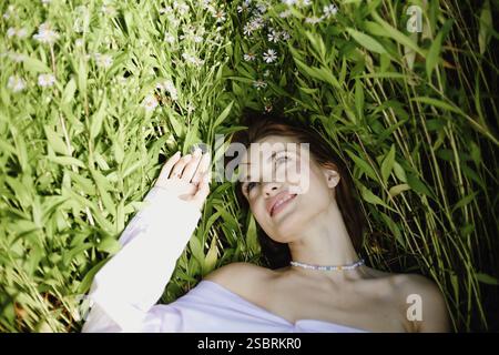 Eine Frau liegt zwischen hohen grünen Pflanzen mit nachdenklichem Ausdruck, umgeben von kleinen weißen Blumen Stockfoto