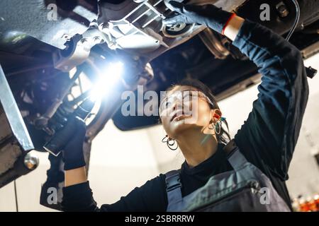 Junge Mechanikerin, die eine Schutzbrille mit Taschenlampe trägt, während sie in einer professionellen Werkstatt das Fahrgestell überprüft Stockfoto