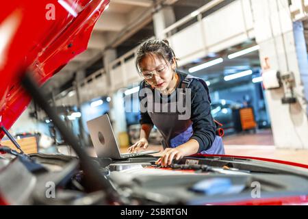 Junge Frau, die als Mechanikerin arbeitet, einen Laptop benutzt, während sie den Motor eines roten Autos in einer viel befahrenen Werkstatt inspiziert Stockfoto