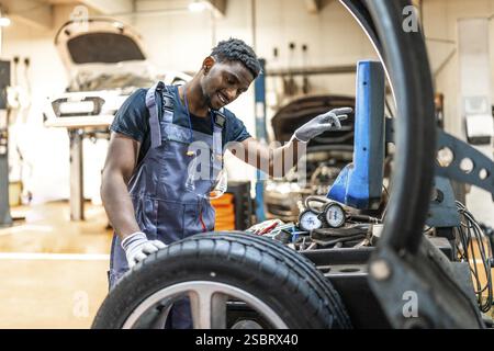 Junger, lächelnder afroamerikanischer Mechaniker, der Autoreifen mit einem digitalen Reifenwechsler in einer Autowerkstatt wechselt Stockfoto