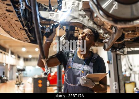 Junger Mechaniker, der in einer modernen Werkstatt mit Taschenlampe und Tablett das Fahrgestell inspiziert Stockfoto